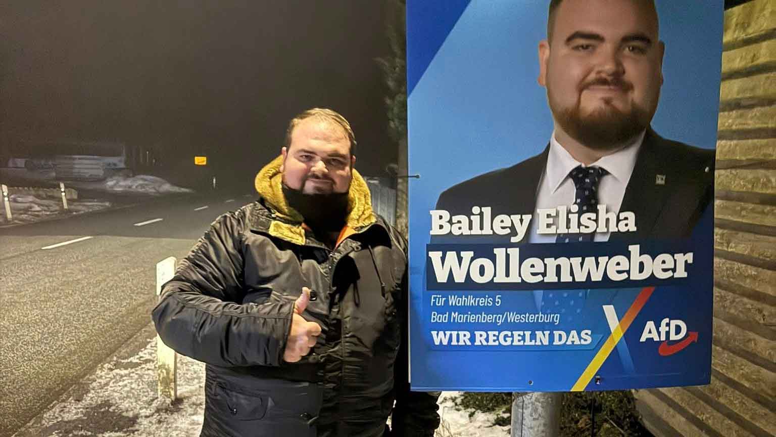 Bailey Wollenweber steht nachts an einer Straße im Winter neben einem Wahlplakat der AfD. Er trägt eine schwarze Jacke mit gelbem Innenfutter und zeigt mit dem Daumen nach oben. Neben ihm hängt ein großes blaues Wahlplakat mit dem Porträt von ihm selbst im Anzug sowie der Schriftzug eines Landtagskandidaten der AfD für den Wahlkreis Bad Marienberg/Westerburg.